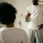 Back view of unrecognizable black boy with Afro hair standing in front of crop anonymous dad pointing finger and scolding at home