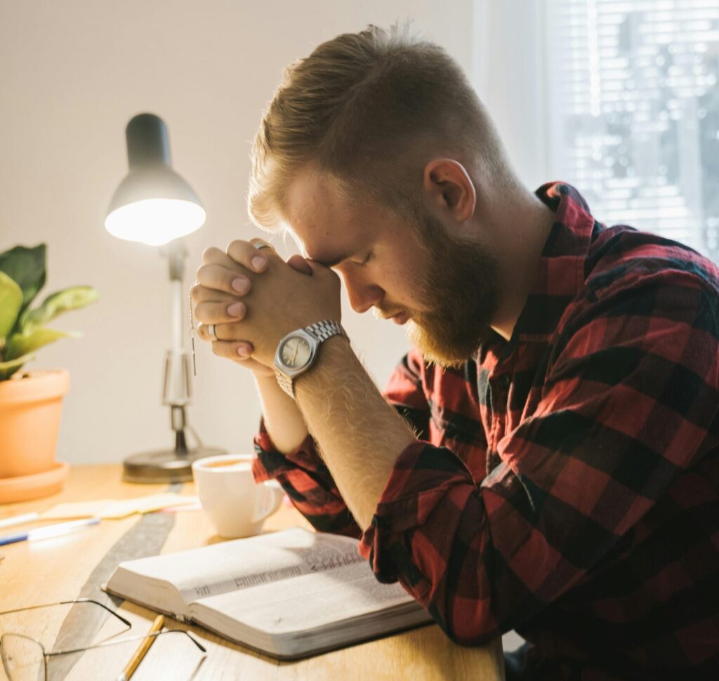 Bearded man in plaid shirt praying with closed eyes at desk with Bible under soft indoor lighting.
