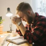 Bearded man in plaid shirt praying with closed eyes at desk with Bible under soft indoor lighting.