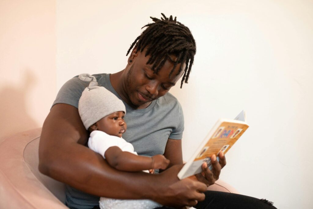 A father with dreadlocks reads a book to his baby, creating a bonding moment.