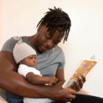 A father with dreadlocks reads a book to his baby, creating a bonding moment.
